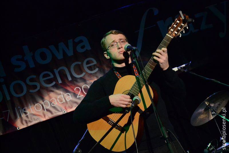 Młody mężczyzna , w okularach, na scenie, gra na gitarze klasycznej.